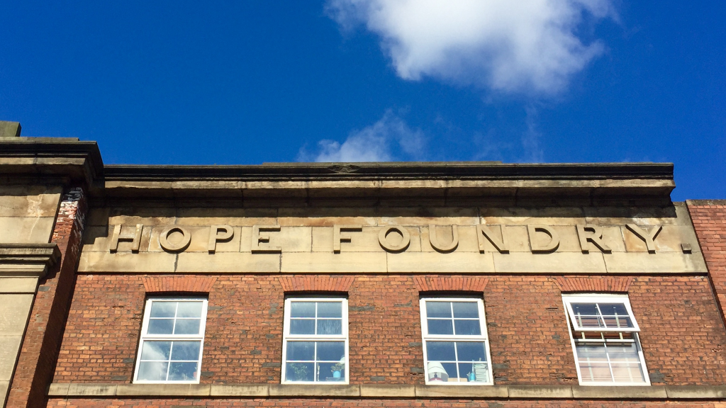 An image taken from the ground looking up to sky which includes the top of the Hope Foundry building. This part of the building has 'Hope Foundry' carved into the side of the building.