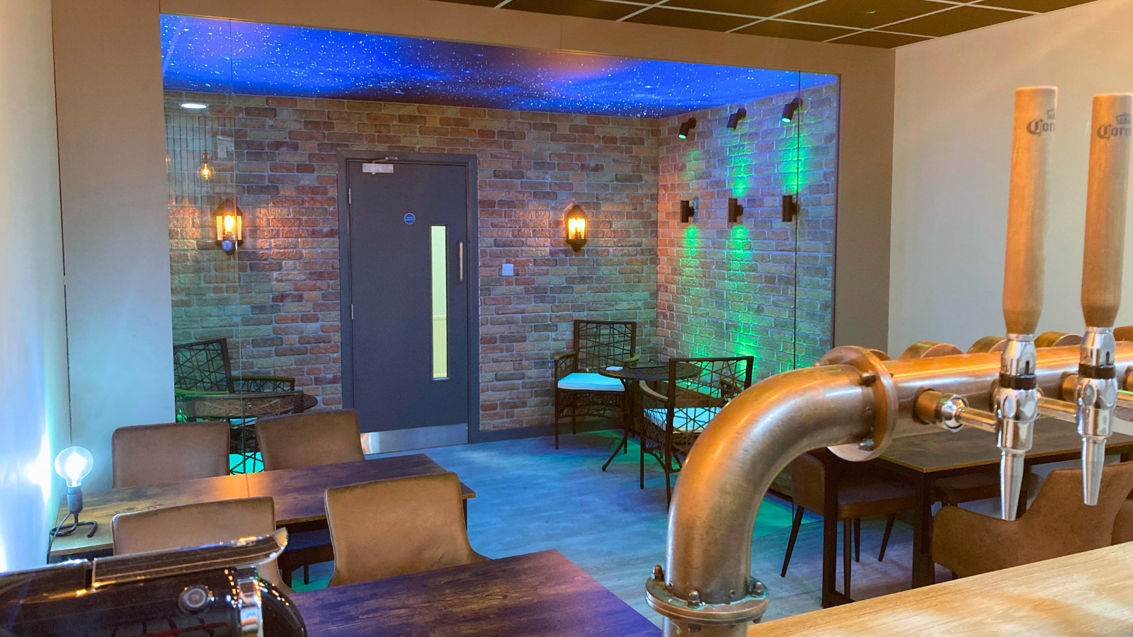 A picture taken from the bar looking over the seating and towards the door. There are a couple of tables with chairs in the shot, there are lamps on the tables with other LED lights displaying blue and green colours. This image is taken from the left hand side of the room.
