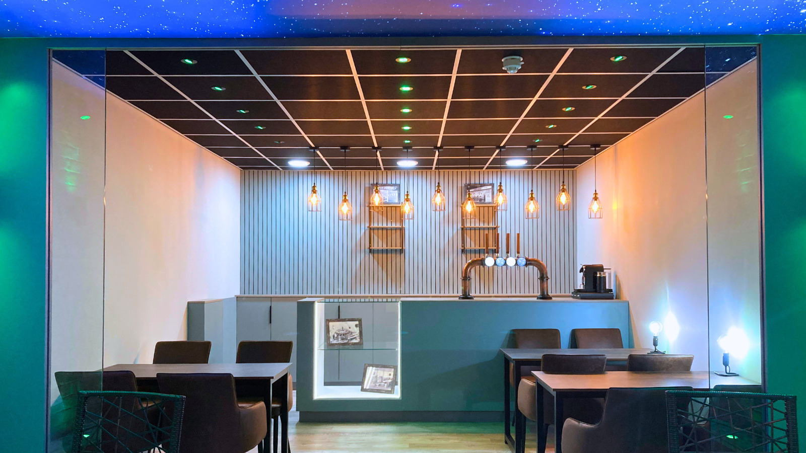 A picture taken from the door looking over the seating and towards the bar. There are a couple of tables with chairs in the shot, there are lamps on the tables with other LED lights displaying blue and green colours. This image is taken from the back of the room in the middle.