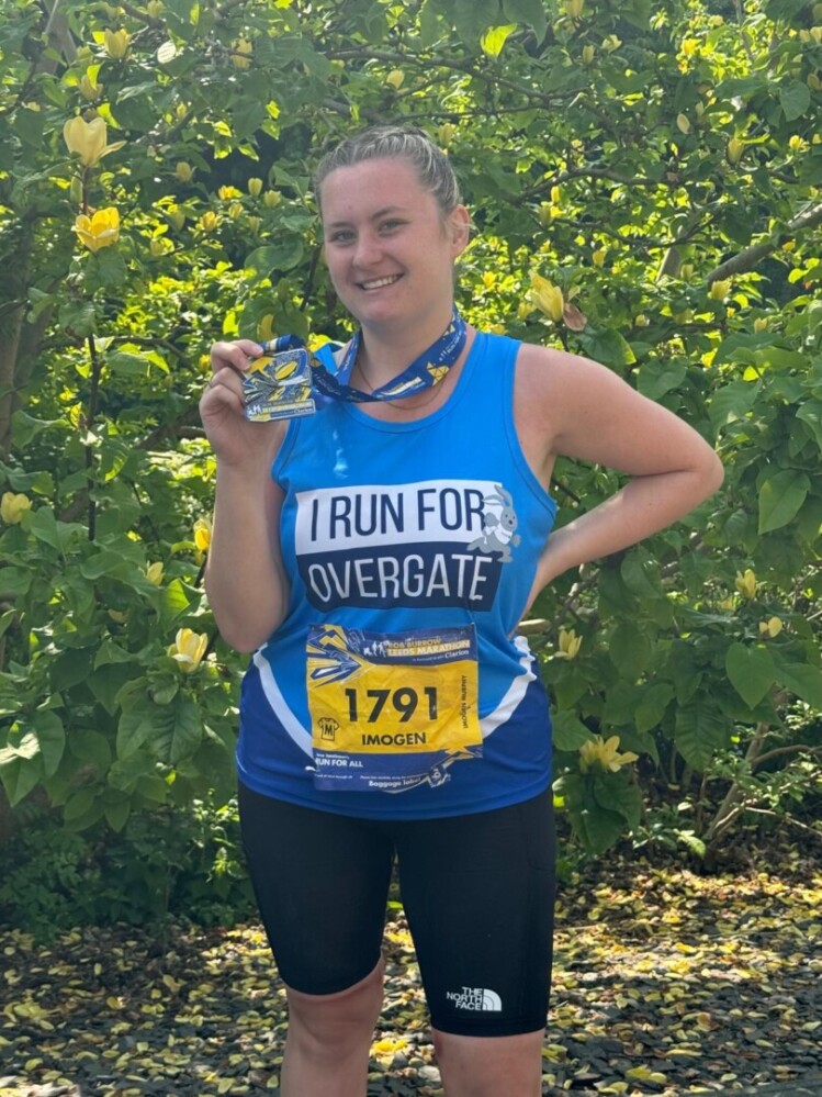An image of Imogen holiday up her medal and running the Rob Burrows Leeds Marathon in 2023 for Overgate.